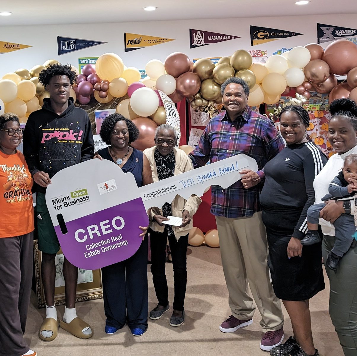 Teen Upward Bound team celebrates closing on their property, surrounded by balloons and college pennants. They smile together, posing with a large novelty key.