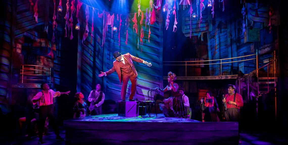 Photo of "A Wonderful World" theatre production by Miami New Drama. The dark stage is lit up by bright purple lights as a man in a red suit floats above the ground.